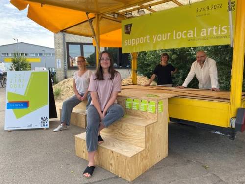 Vier Personen posieren, umgeben von Werbeplakaten zum ZINNOBER, an einem überdachten, mobilen Infostand.